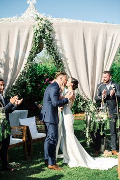 a bride and groom kiss as they stand in front of an outdoor wedding ceremony arch