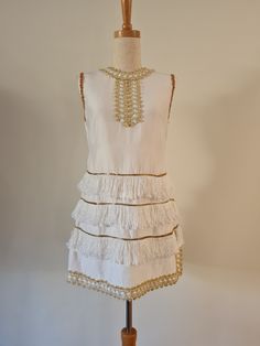 A Gown of Beauty fashioned by Jeanne De Camp Melbourne White and Gold 1960's mini dress/tunic with rows of fringing detail and side splits. Sleeveless with a crew neck, this has metallic braid detailing at the neck and armholes.  The Gold and White braided detail goes all around the neck, and along the hem edge and side seams, with a separate sequin braid around the armholes.  There are three rows of braided detail on the lower hem each with a gold lurex detail at the top edge.  Fully lined this White Sleeveless Flapper Dress, White Sleeveless Mini Dress With Fringe, Gold Sleeveless Mini Dress With Contrast Sequin, Gold Braids, 1960s White Mini Dress, 1960s Mini Dress, Evening V-neck Mini Dress With Fringe, Gogo Dress, Camp Camp