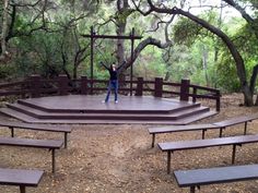 a woman standing on top of a wooden stage surrounded by benches and trees in the woods