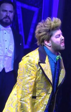 a man in a yellow jacket singing on stage with other men standing behind him and wearing tuxedos