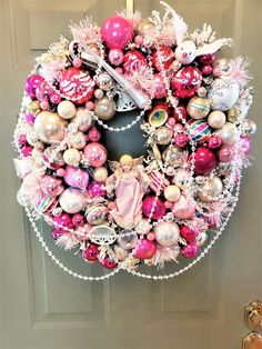 a pink and white christmas wreath hanging on the front door with ornaments all over it