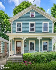 a blue house with white trim on the front and side of it, surrounded by greenery