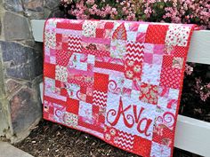 a red and white quilt sitting on top of a wooden bench next to flowers in front of a stone wall