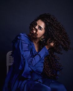 a woman with curly hair sitting on a chair in a blue dress and gold hoop earrings