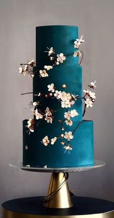 a blue cake with white flowers on the top is sitting on a gold plated stand