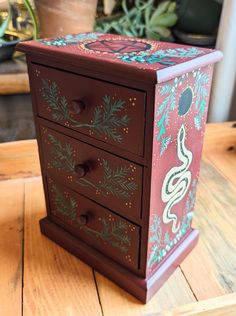 a painted wooden box with drawers on top of a wood table next to potted plants