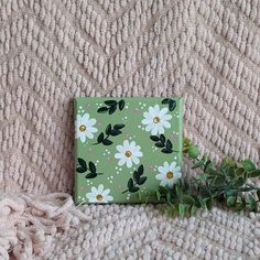 a small green box with white flowers on it sitting on a blanket next to a plant