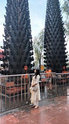 a woman standing next to a metal fence in front of tall black trees that have been carved into them