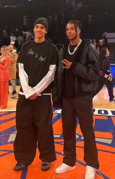 two young men standing next to each other on a basketball court with people in the background