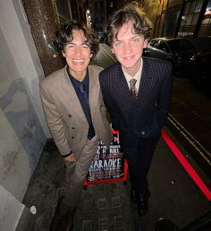 two young men dressed in suits and ties standing next to each other on the sidewalk