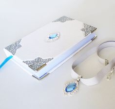 a white book with a blue ribbon around it next to a pair of earrings and a lanyard