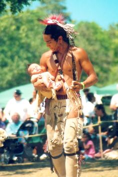 a native american man holding a baby in his arms while people watch from the sidelines