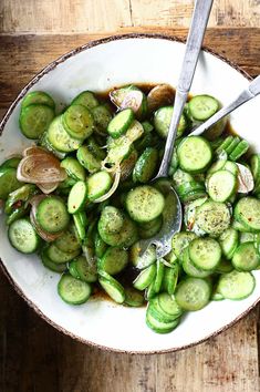 a white bowl filled with sliced cucumbers next to two spoons on top of a wooden table