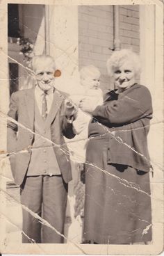 an old black and white photo of two people holding a baby in front of a house