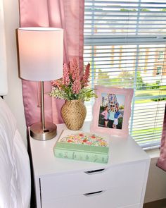 a bedroom with pink curtains and flowers on the nightstand next to a lamp, photo frame and flower pot