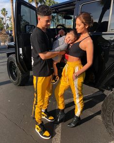 a man standing next to a woman holding a baby in front of a black jeep