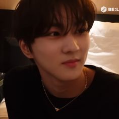 a young man with black shirt and necklace on sitting in front of a white bed