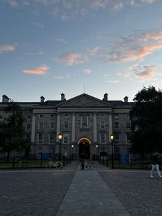 people walking in front of an old building at dusk with the sun setting behind them