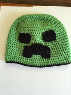 a crocheted green hat with black eyes and a frown on it's face