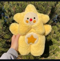 a hand holding a yellow teddy bear in front of a pine tree