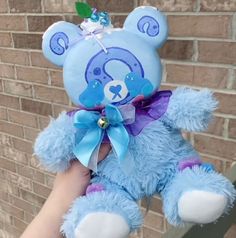 a blue teddy bear with a purple ribbon around it's neck sitting in front of a brick wall