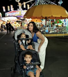 a woman standing next to two small children in a stroller at an amusement park