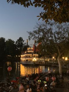 many people are sitting on benches near the water at night with a boat in the background