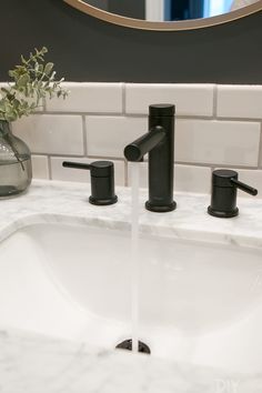 a bathroom sink with two black faucets and a white marble counter top in front of a round mirror