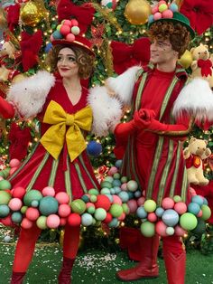 two people dressed in christmas costumes standing next to a christmas tree with decorations on it