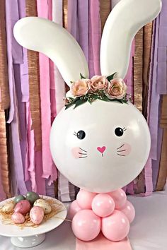 a balloon bunny with flowers on its head sitting next to a plate of cookies and pastries