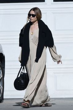 a woman in a long dress and black jacket walking past a white garage door with a handbag