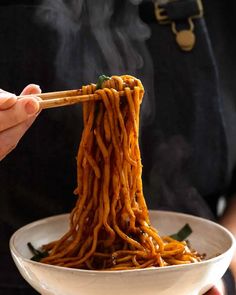 a person holding chopsticks over a bowl of noodles with sauce on it and steam rising from the top