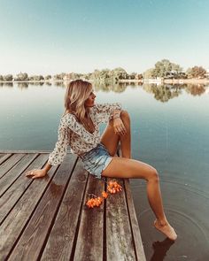 a woman sitting on top of a wooden dock next to the water with her legs crossed