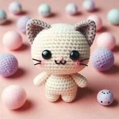 a small crocheted cat sitting next to some eggs on a pink surface with balls in the background