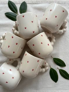 five white cups with red hearts on them sitting on a tablecloth next to green leaves