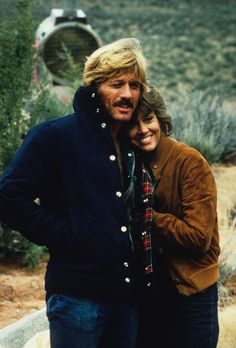 a man and woman standing next to each other in front of a desert area with trees