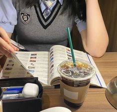 a woman sitting at a table reading a book and holding a drink in her hand