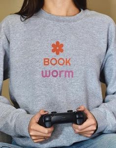 a woman holding a game controller in her hands while wearing a book worm sweatshirt with the words book worm on it