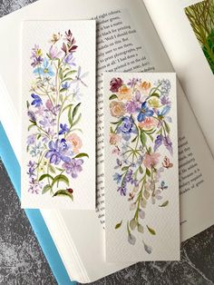 three watercolor cards with flowers on them sitting next to an open book and plant