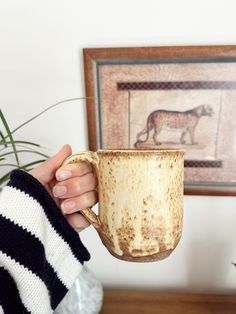 a person holding a coffee cup in their hand with a painting on the wall behind them