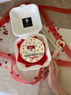 a person holding a cake in a box on a table with red and white confetti