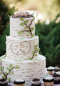 a wedding cake and cupcakes are on a table with trees in the background