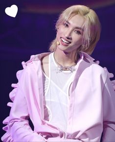 a woman with blonde hair wearing a pink jacket and smiling at the camera while standing in front of a purple background