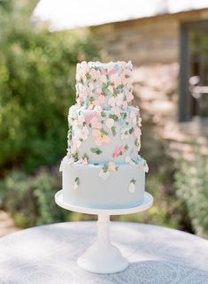 a three tiered cake with pink flowers on it sitting on a white table outside