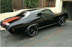 a black muscle car parked in front of a garage with an orange stripe on the hood