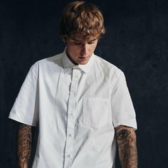 a man with tattoos standing in front of a black background wearing a white shirt and jeans