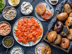 an assortment of different foods on plates and in bowls next to breads, bagels, dips