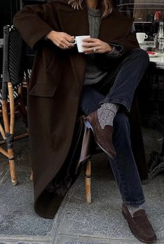 a woman sitting in a chair holding a cup and looking at the camera while wearing a brown coat