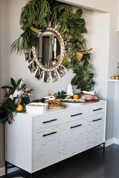 a white dresser topped with lots of greenery next to a wall mounted round mirror