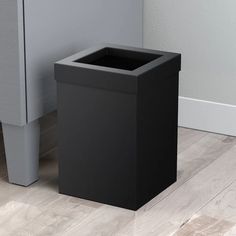 a black trash can sitting on the floor next to a white wall and wooden floors